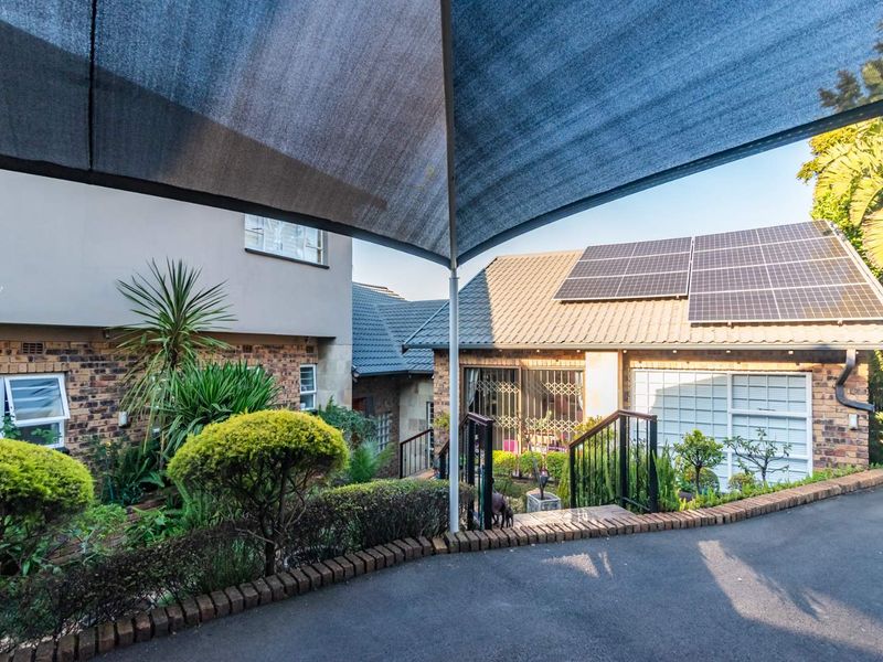 Front exterior view featuring a paved driveway, established garden, shade sail structure, and roof-mounted solar panels on the main dwelling.
