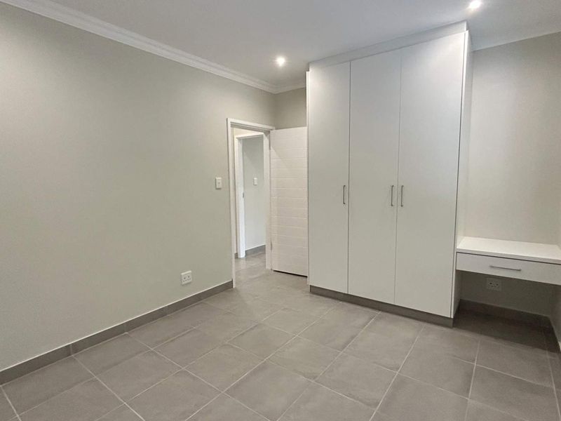 A clean, modern bedroom featuring neutral tiled flooring, built-in white cupboards, and an integrated desk area with recessed lighting.