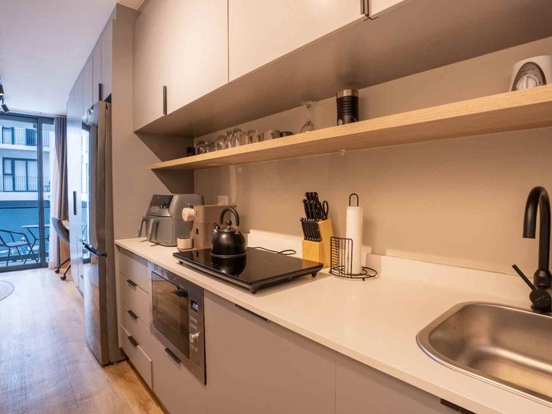 Modern galley-style kitchen featuring sleek grey cabinetry, stone countertops, integrated appliances, and a floating timber shelf, leading to a balcony.