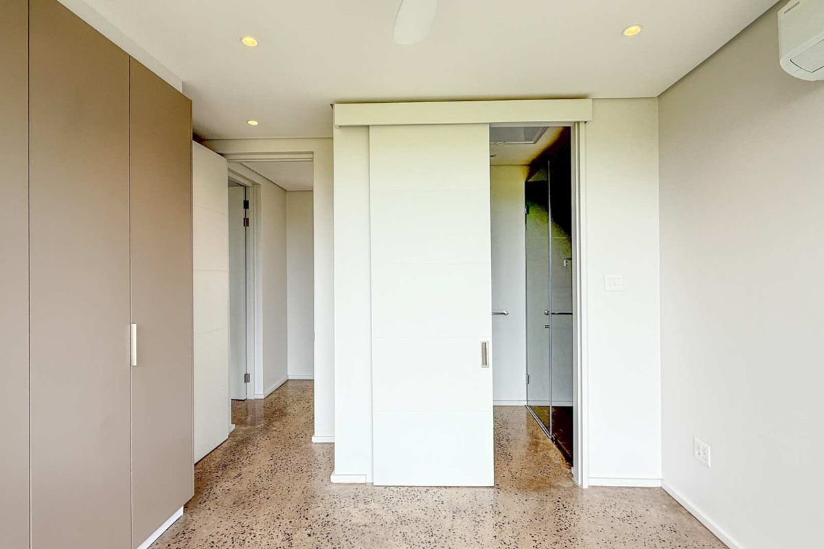 A modern bedroom interior featuring polished concrete flooring, built-in floor-to-ceiling cabinetry, a sliding door leading to an en-suite bathroom, and a wall-mounted air conditioning unit.