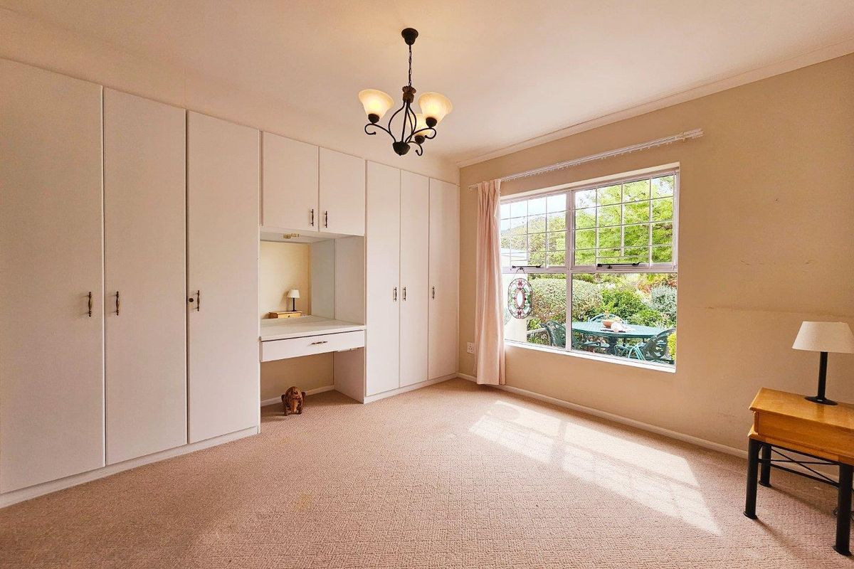A well-lit bedroom featuring extensive built-in cabinetry with an integrated vanity desk, carpeted flooring, and a large window overlooking the garden.