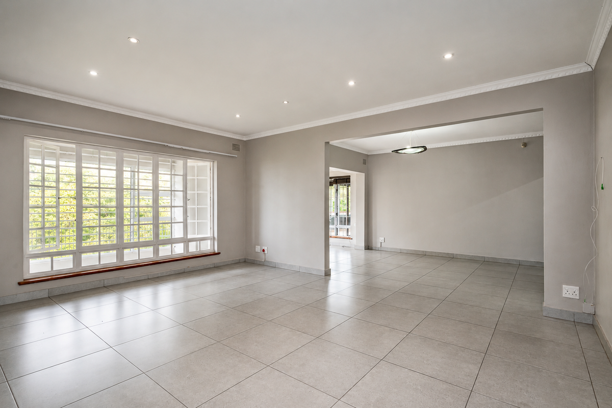 A spacious, light-filled living area featuring neutral tiled flooring, recessed downlighting, and a large bay window with security bars.