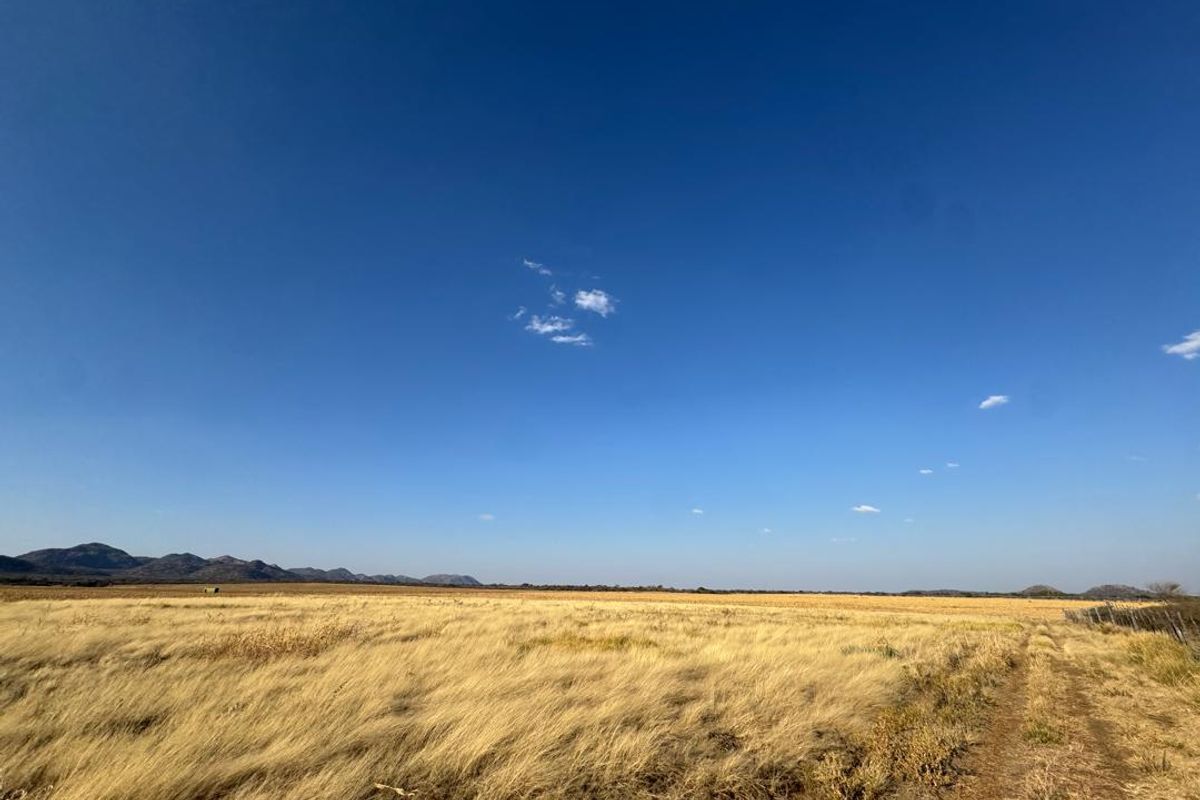 Expansive open farm landscape featuring dry grassland, distant mountain ranges, and a clear blue sky.