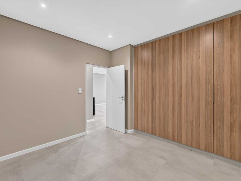 A clean, modern bedroom featuring neutral wall tones, large-format floor tiles, and extensive built-in wooden cabinetry.