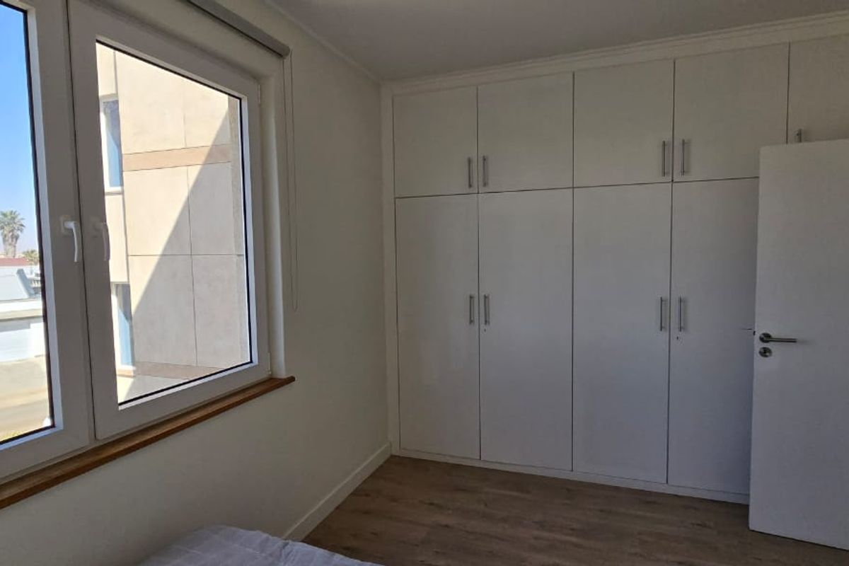A bright, empty bedroom featuring modern white built-in cupboards, laminate flooring, and large windows offering a clear exterior view.