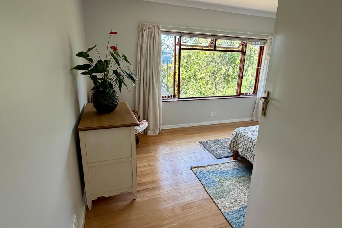 A bright bedroom featuring polished hardwood flooring, crown molding, and large windows with decorative security bars overlooking greenery.