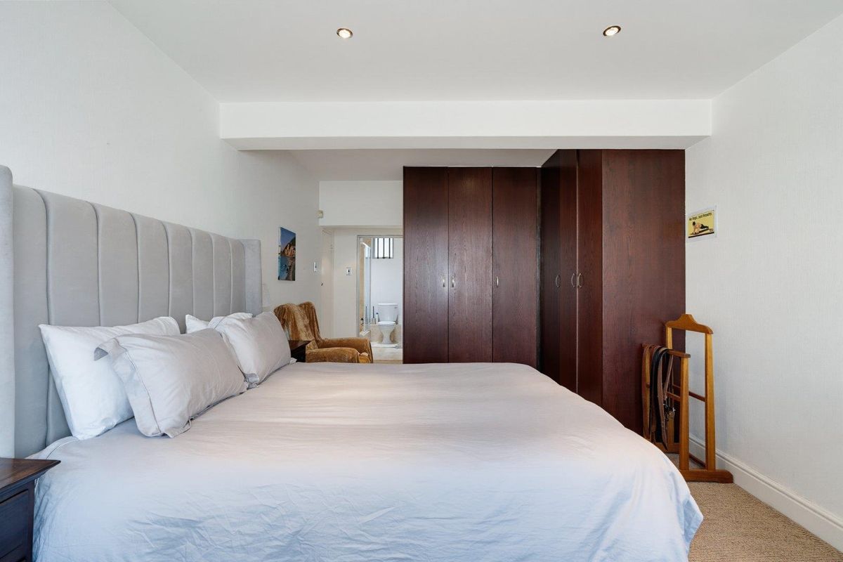 A well-maintained bedroom featuring built-in dark wood cupboards, recessed ceiling lighting, and neutral carpeting, with an en-suite bathroom visible in the background.