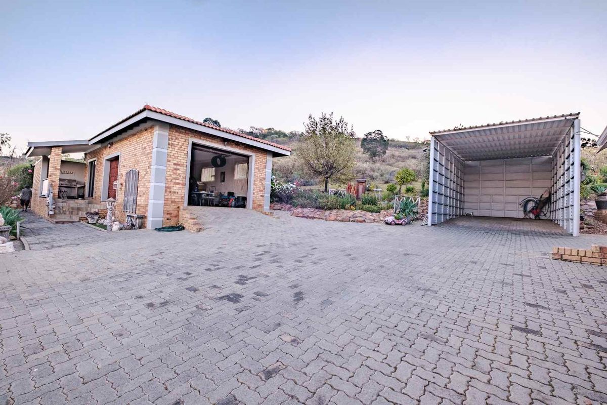 Exterior view featuring a brick garage, a separate metal carport, and a paved driveway area set against a natural hillside landscape.