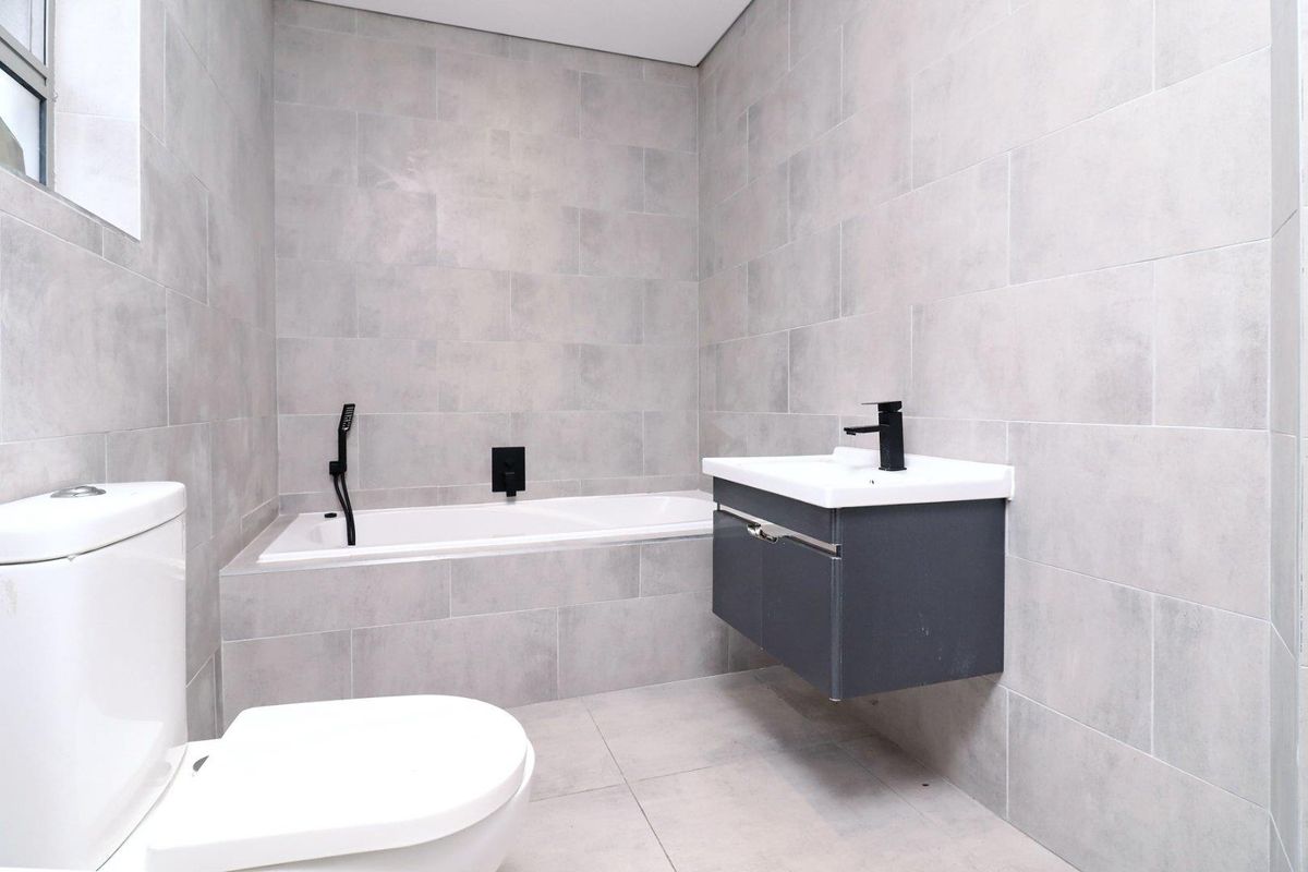 Modern bathroom featuring grey tiled walls and flooring, a wall-mounted vanity with a basin, and a built-in bathtub with black fixtures.