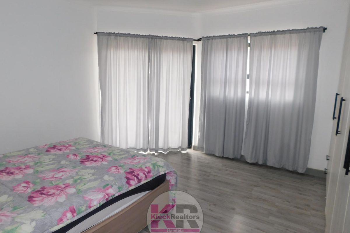 A bright bedroom featuring light-toned wood-look flooring, neutral walls, and large windows dressed with sheer curtains.