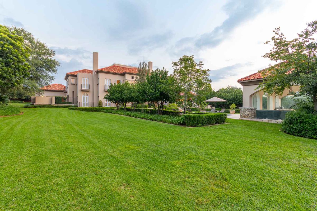 Expansive rear garden featuring manicured lawns, mature landscaping, and a view of the main residence and outdoor entertainment structure with terracotta roof tiles.