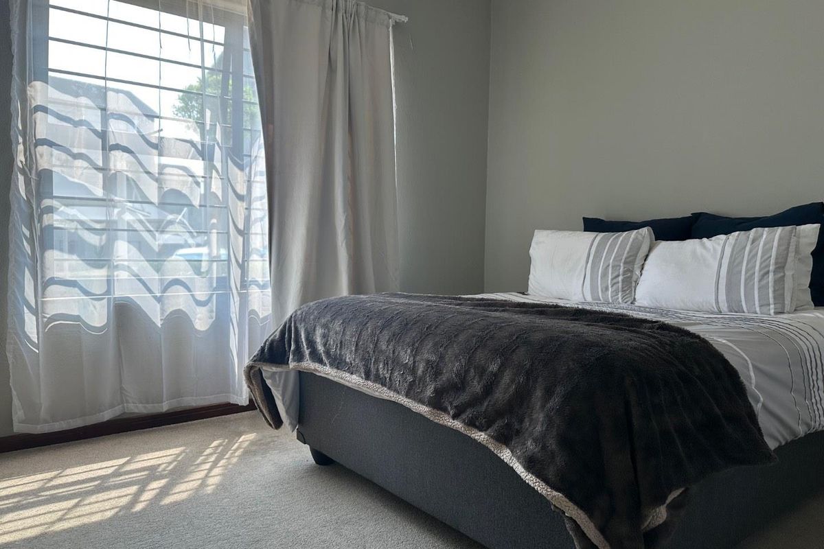 A well-lit bedroom featuring neutral wall tones, carpeted flooring, and a large window with natural light access.