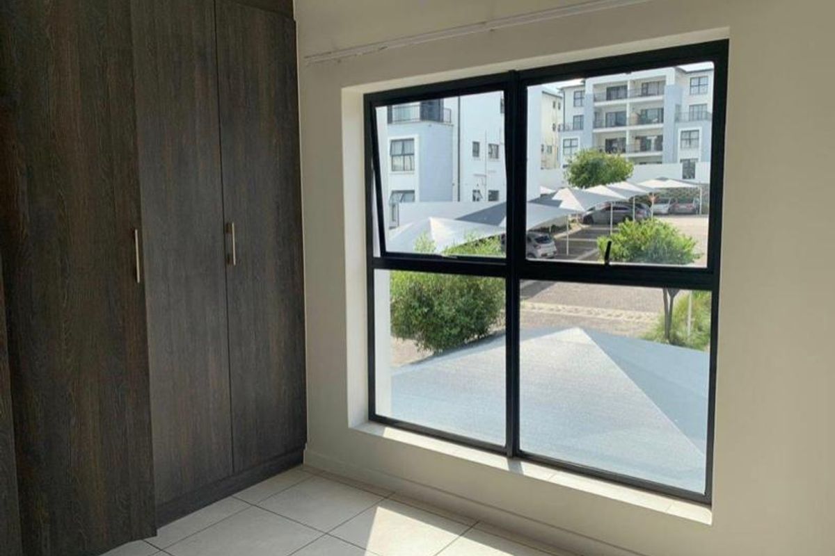A bright, empty bedroom featuring modern dark wood built-in cupboards, neutral tiled flooring, and large aluminium-framed windows overlooking the complex parking area.