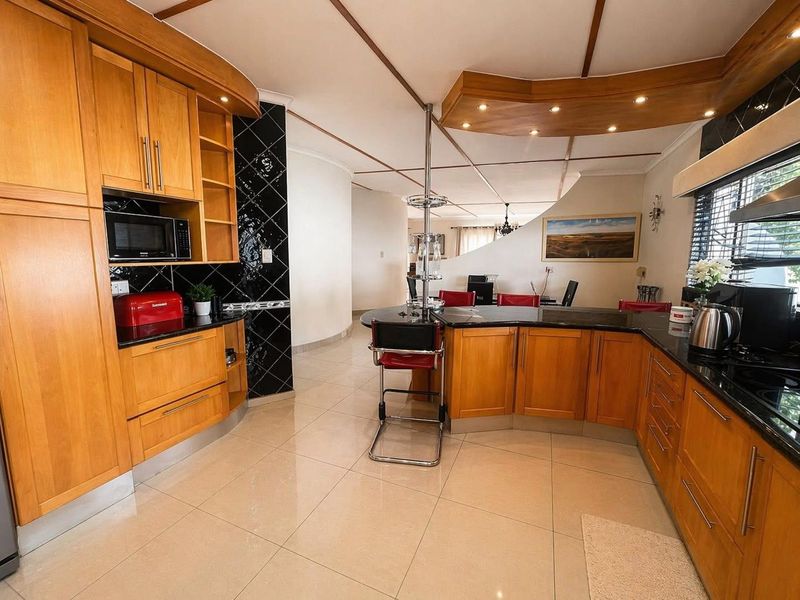 A spacious kitchen featuring warm wooden cabinetry, black granite countertops, a breakfast bar, and modern tiled flooring with integrated recessed lighting.