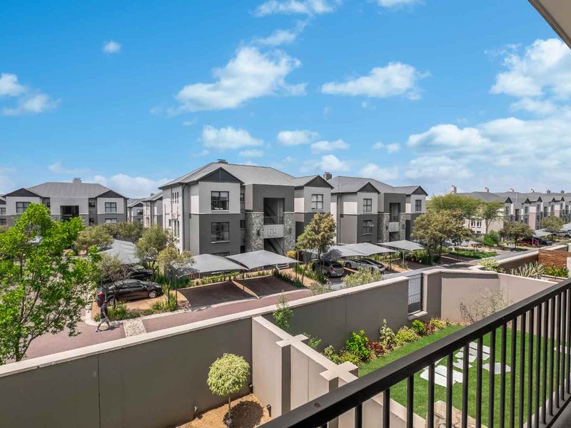 Elevated view from a balcony overlooking the apartment complex, featuring landscaped grounds, covered parking bays, and modern residential building architecture.