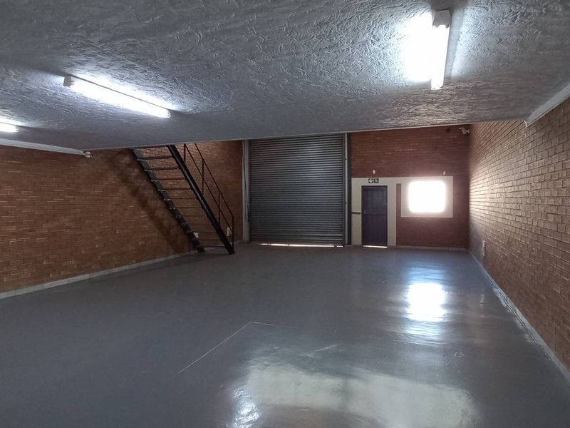 Spacious industrial warehouse unit featuring exposed brick walls, polished concrete flooring, a large roller shutter door, and a metal staircase leading to an upper level.