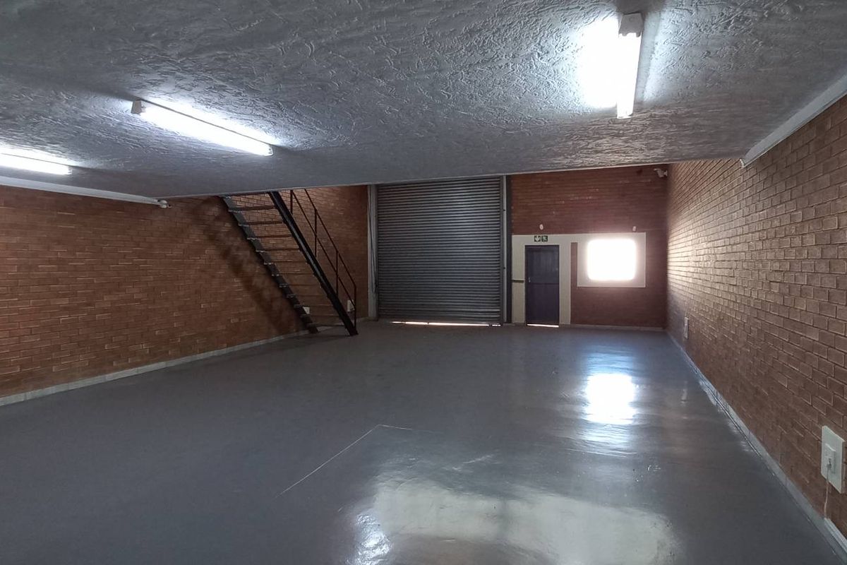 Spacious industrial warehouse unit featuring exposed brick walls, polished concrete flooring, a large roller shutter door, and a metal staircase leading to an upper level.
