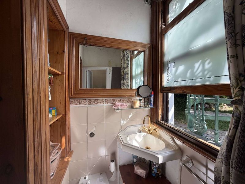 A compact bathroom featuring a corner vanity basin with brass fixtures, built-in wooden shelving, and a framed mirror, requiring cosmetic updates.