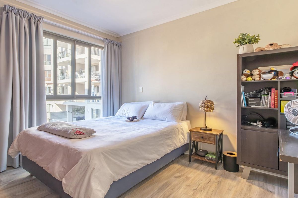 A well-lit bedroom featuring light-toned laminate flooring and a large sliding window with neutral curtains, offering a comfortable and functional living space.