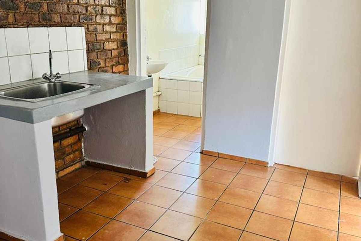 A functional kitchen area featuring exposed brick walls, tiled flooring, and a concrete counter with a stainless steel sink, leading into a bathroom.