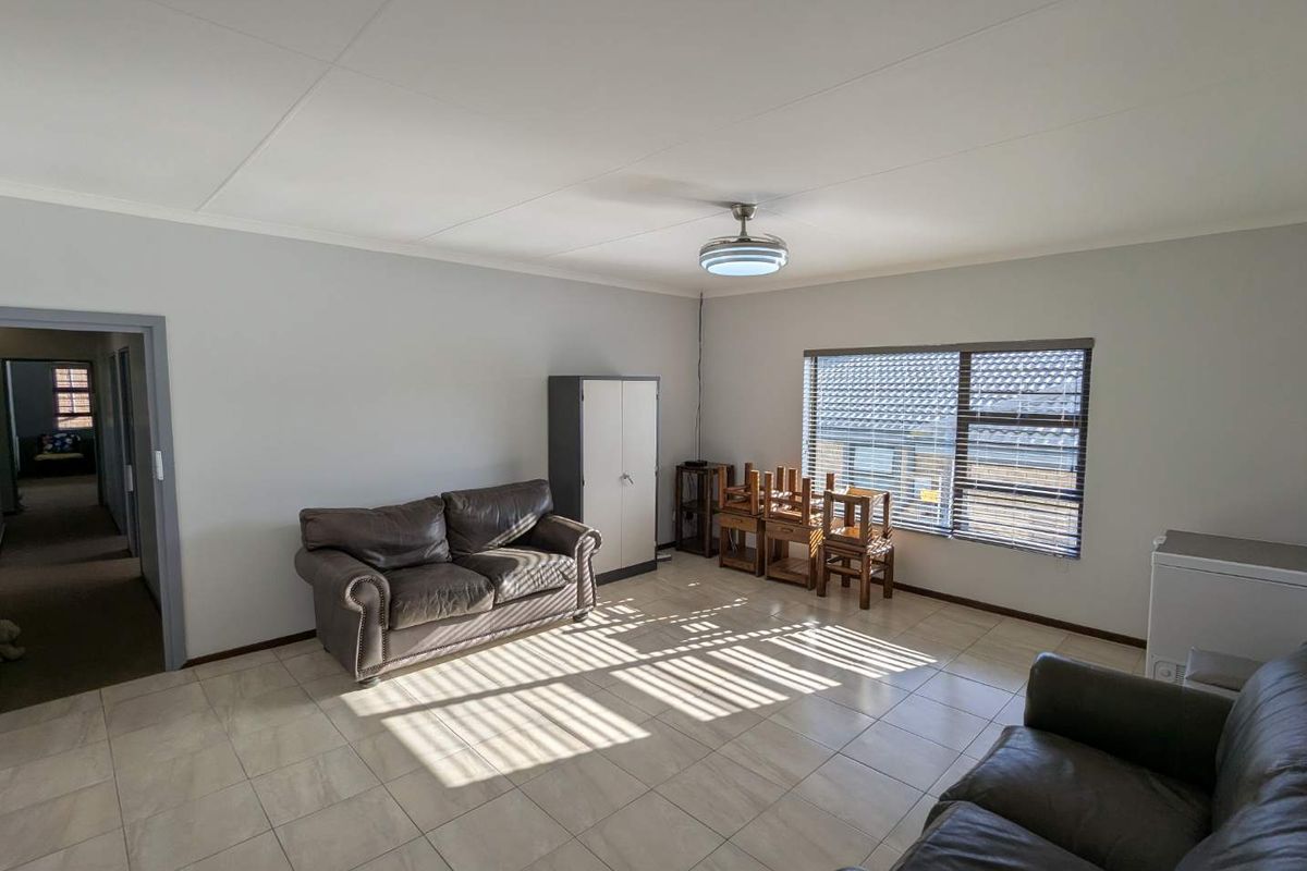 A spacious living room featuring neutral tiled flooring, a ceiling fan, and large windows with venetian blinds, offering a bright and functional living space.