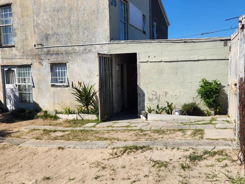 Rear exterior view of the property showing a weathered facade, security features, and an unpaved garden area requiring landscaping.