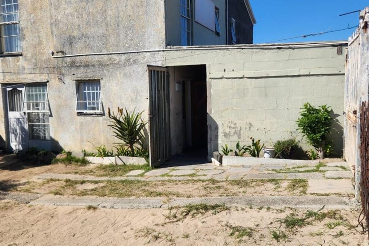 Rear exterior view of the property showing a weathered facade, security features, and an unpaved garden area requiring landscaping.