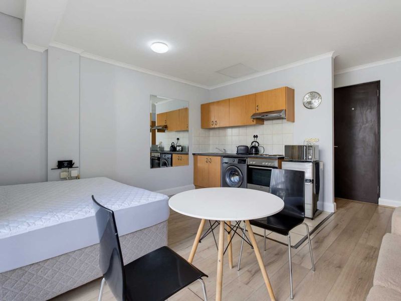 Compact open-plan studio apartment featuring a functional kitchenette with built-in cabinetry, oven, and hob, finished with light laminate flooring.