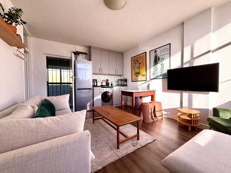 Open-plan living area featuring a compact kitchen with built-in cabinetry, tiled backsplash, and laminate flooring, leading to a secure exterior entrance.