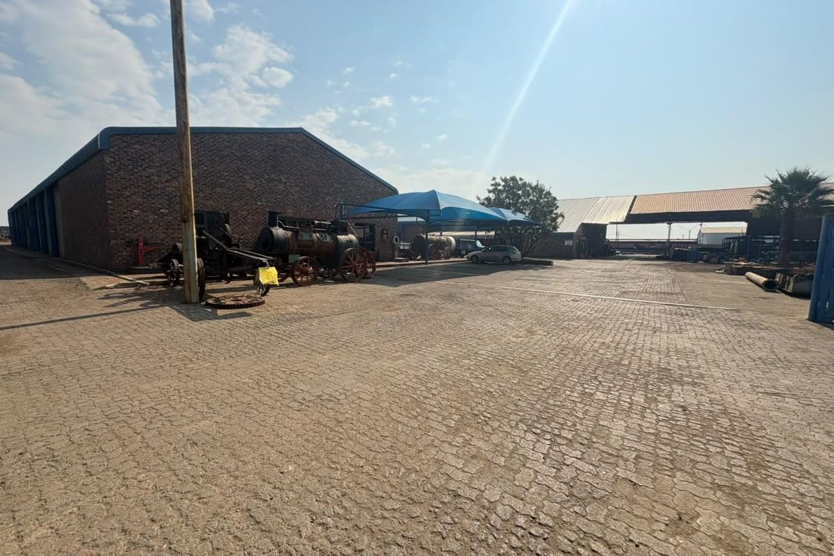 Spacious industrial yard featuring a brick warehouse, paved surface, and covered parking structures suitable for various commercial or storage operations.