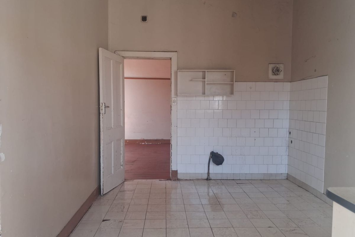 An empty, dated kitchen area featuring tiled flooring, a tiled splashback, and wall-mounted shelving, requiring renovation to modernize the space.