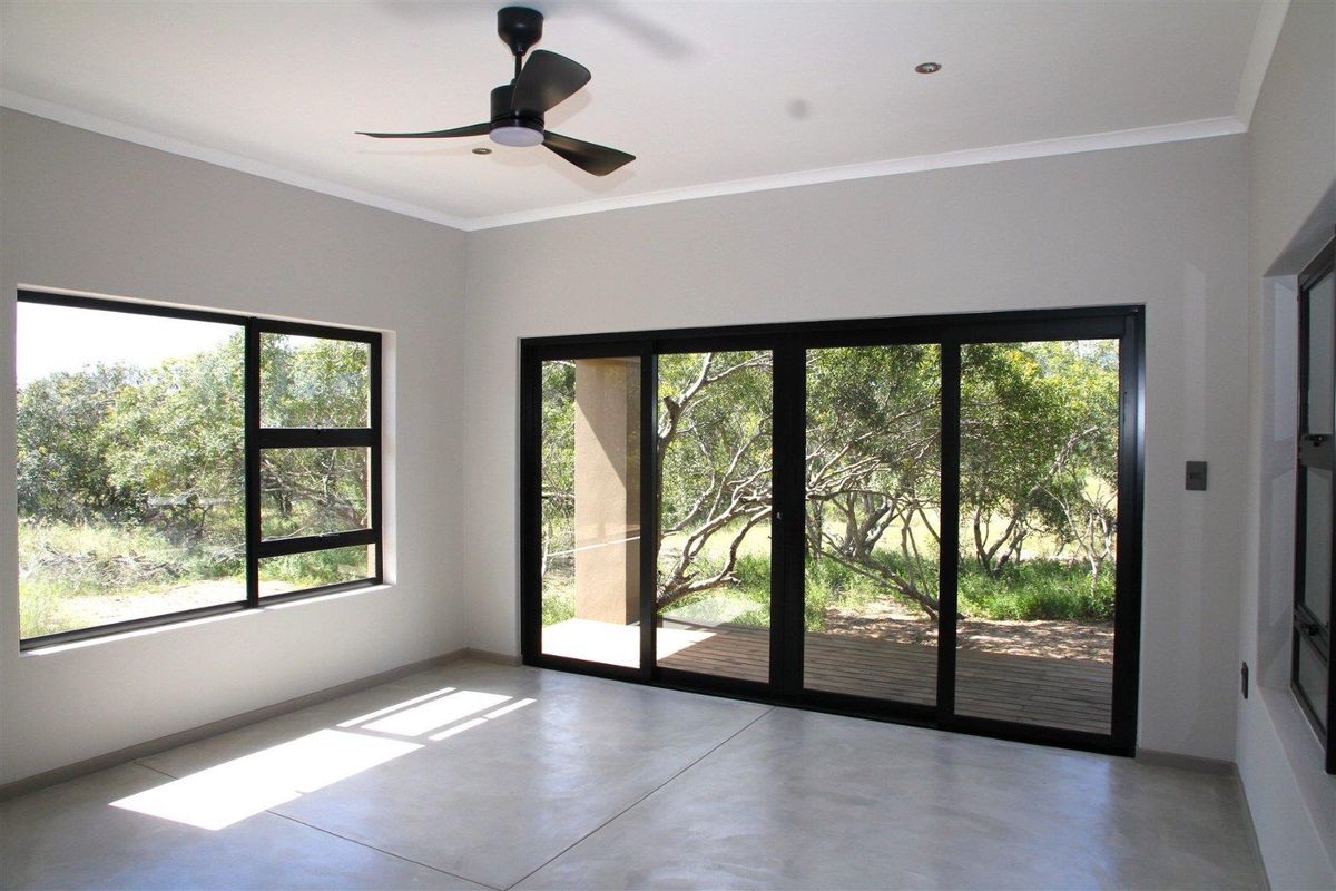 A bright, modern living room featuring polished concrete floors, a ceiling fan, and large black-framed sliding doors opening onto a wooden deck.