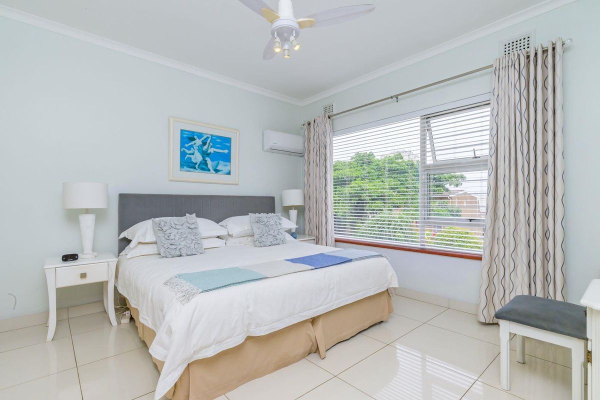A bright bedroom featuring tiled flooring, a ceiling fan, and a wall-mounted air conditioning unit, with large windows providing natural light.