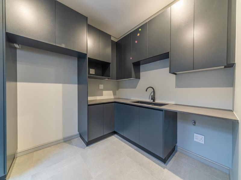 A modern, clean scullery featuring dark grey built-in cabinetry, a sleek undermount sink with a black mixer tap, and neutral tiled flooring.