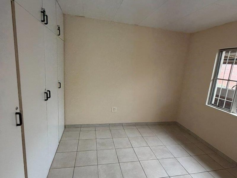 A simple, empty bedroom featuring tiled flooring, built-in cupboards, and a window with security bars, offering a clean canvas for potential updates.