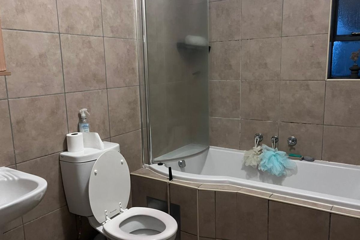 A functional bathroom featuring a pedestal basin, toilet, and a built-in bathtub with a glass shower screen, finished with neutral wall tiling.