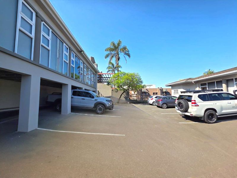 Exterior view of the property featuring a paved parking area with covered bays and multi-pane windows along the building facade.