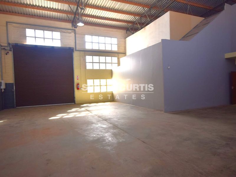 Spacious industrial warehouse unit featuring high ceilings, a large roller shutter door, and ample natural light through industrial-style windows.
