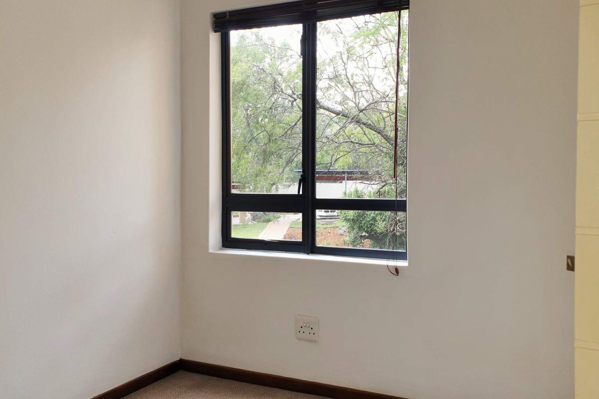 A bright, empty bedroom featuring neutral walls, carpeted flooring, and a window with dark framing overlooking the exterior.