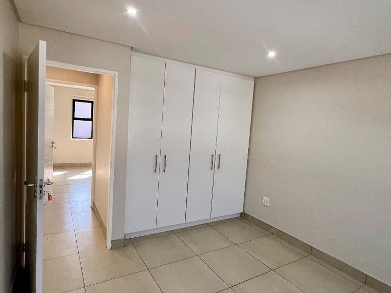A clean, empty bedroom featuring neutral tiled flooring, built-in white cupboards, and recessed downlighting, offering a bright and versatile living space.