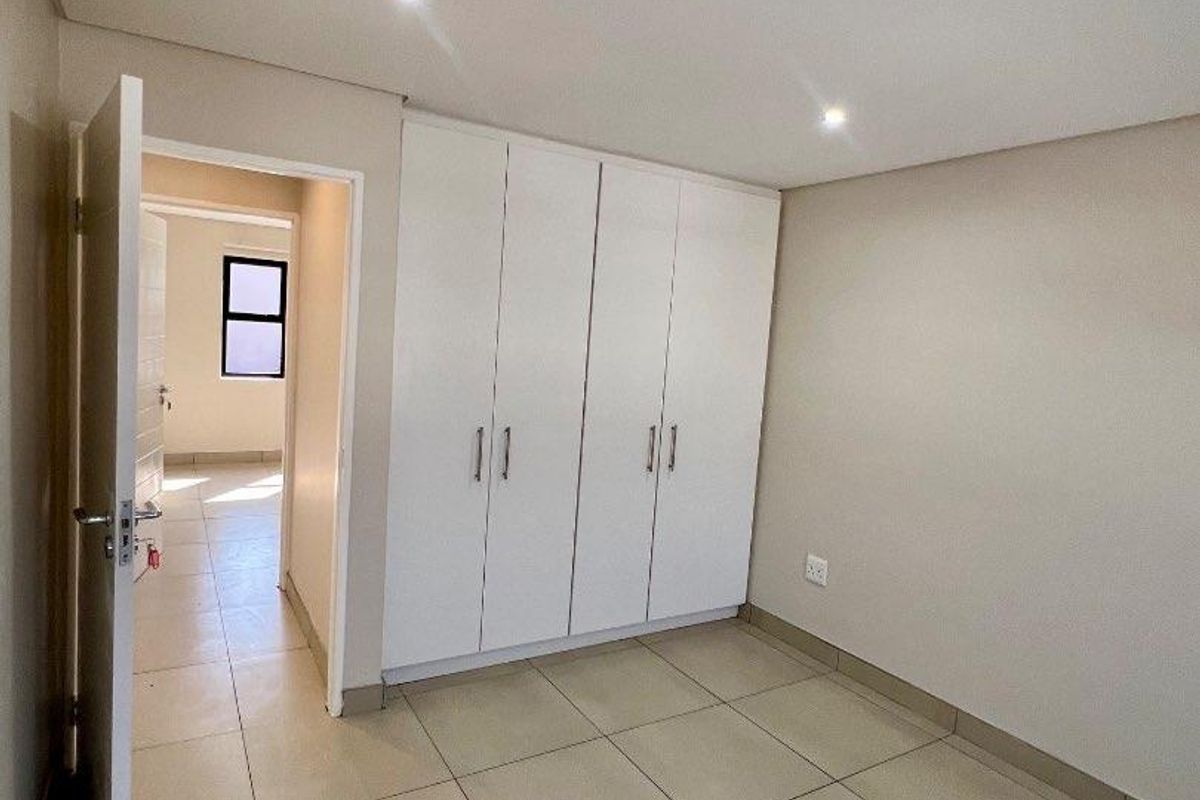 A clean, empty bedroom featuring neutral tiled flooring, built-in white cupboards, and recessed downlighting, offering a bright and versatile living space.