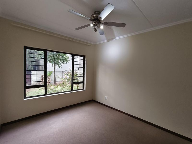 A clean, empty bedroom featuring neutral walls, carpeted flooring, a ceiling fan, and a large window with security bars overlooking the garden.