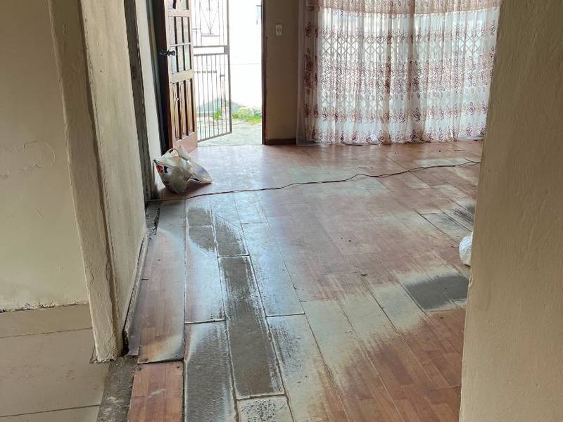An interior living area currently undergoing renovation, featuring exposed ceiling joists, worn laminate flooring, and a security gate leading to the exterior.