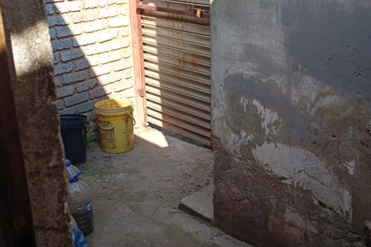 A narrow exterior rear passage featuring a brick boundary wall, concrete flooring, and a metal security gate showing signs of wear and weathering.