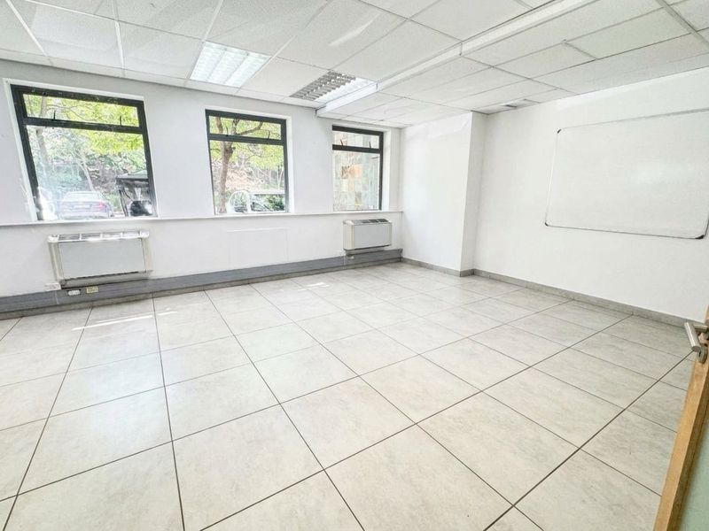 A bright, empty office space featuring tiled flooring, suspended ceilings with recessed lighting, large windows, and wall-mounted HVAC units.