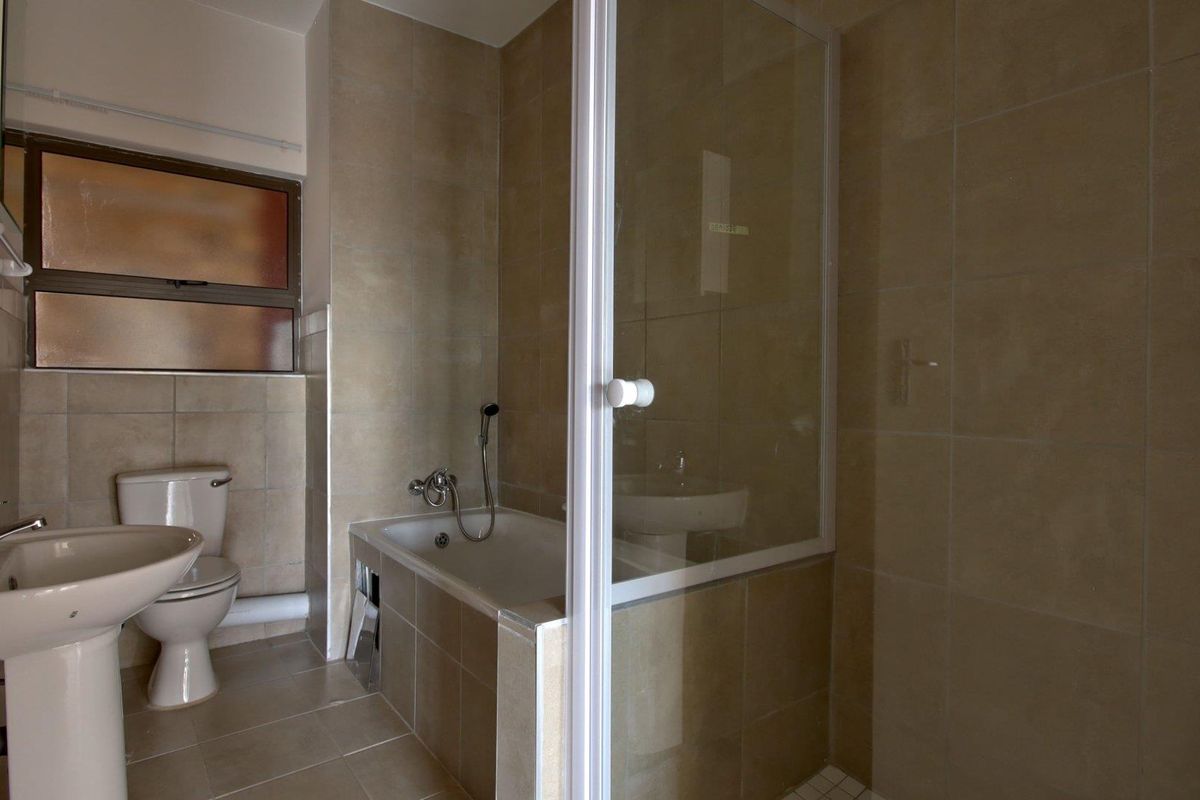 A functional bathroom featuring a pedestal basin, toilet, and a combined bathtub with a shower screen, finished with neutral wall and floor tiling.