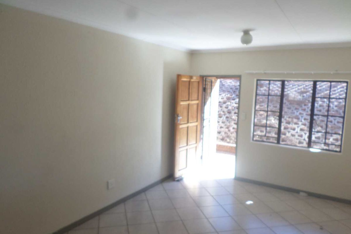 An empty living room featuring tiled flooring, a wooden entrance door, and windows fitted with security burglar bars.