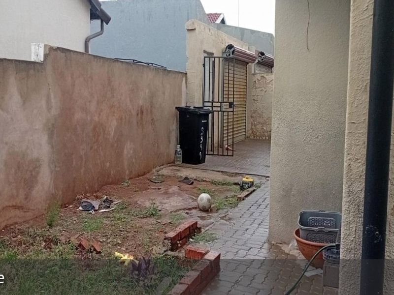 A rear exterior area featuring a paved walkway, boundary wall, and access to a garage with a roller shutter door, requiring maintenance and landscaping.
