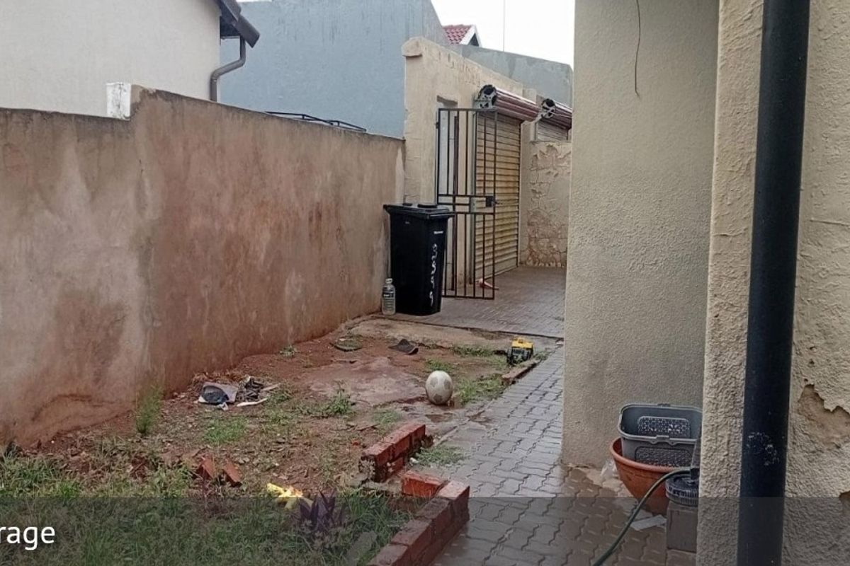 A rear exterior area featuring a paved walkway, boundary wall, and access to a garage with a roller shutter door, requiring maintenance and landscaping.