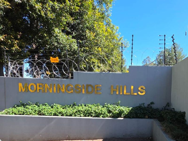 Entrance signage for Morningside Hills featuring a concrete retaining wall, paved driveway, and security electric fencing.